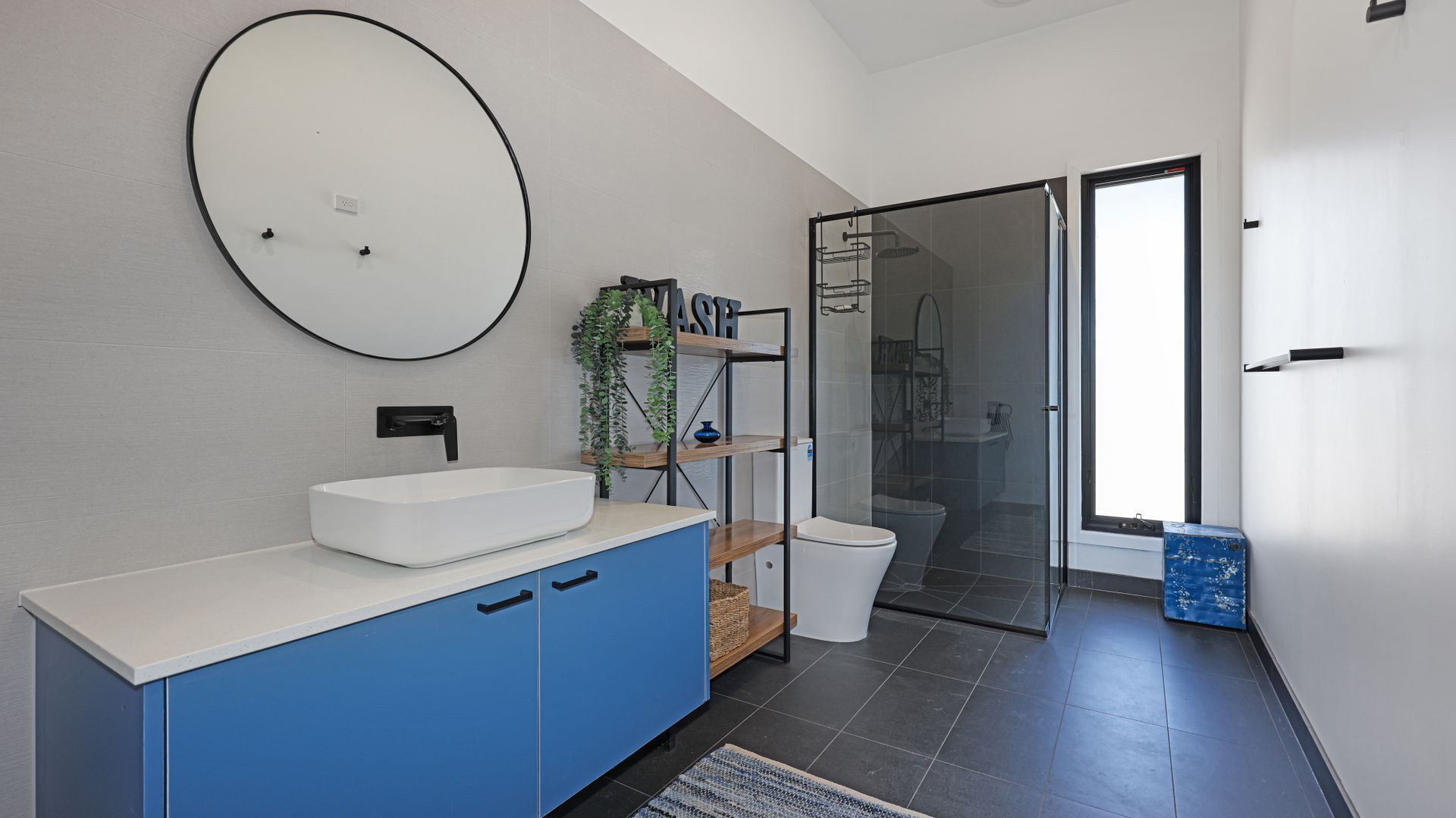 A modern bathroom with a white sink, blue cabinets, a round mirror, and a glass shower enclosure.