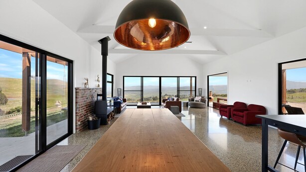 Uma sala de estar espaçosa com piso de madeira prolongado, uma luminária pendente preta grande e vista para o campo através de amplas janelas.