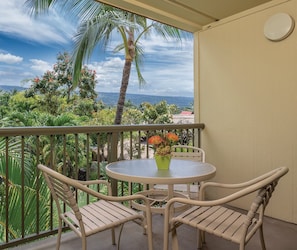 A balcony with a round table and two chairs, overlooking a lush garden and a view of the ocean. - Beautiful 2 bedroom Condo @ Kona! (Kailua-Kona)