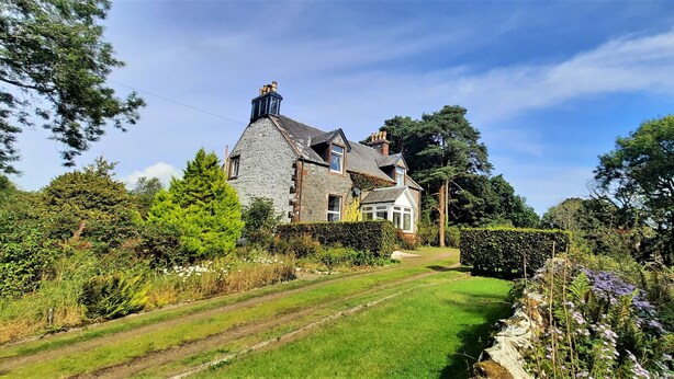 A large stone house with a steeply pitched roof, surrounded by lush greenery and a well-maintained lawn.