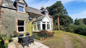 A stone house with a conservatory, a garden, and a paved patio with chairs.