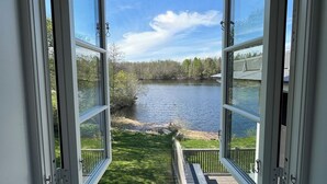Vista desde una ventana abierta hacia un lago, un área con césped y una terraza de madera.