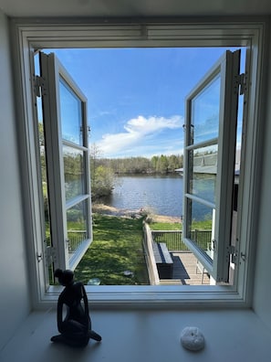 Vista desde una ventana abierta hacia un lago, un área con césped y una terraza de madera.