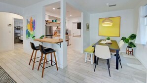 Una cocina moderna con armarios blancos, un refrigerador de acero inoxidable y una encimera de madera. Hay dos taburetes de bar y una mesa de comedor con sillas.