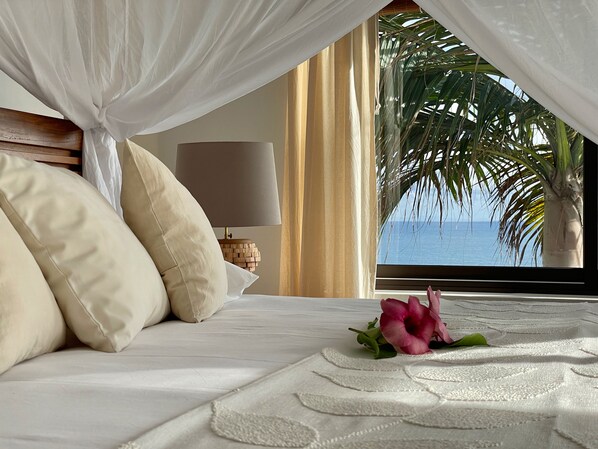 A bedroom with a bed, pillows, a bedside lamp, a flower, and a view of the ocean through a window. - Chalets Bougainville
Upper floor Apartment with Ocean View (Takamaka)