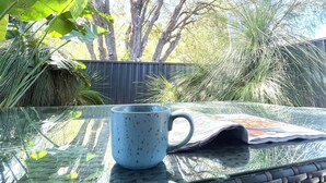 A glass table with a blue mug and a newspaper. Outside, there are trees and a fence. - Quedji Cottage - Beautiful 2 bed private cottage 3 min walk to Dunsborough town. (Dunsborough)