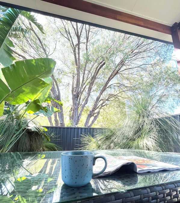 A glass table with a blue mug and a newspaper. Outside, there are trees and a fence.