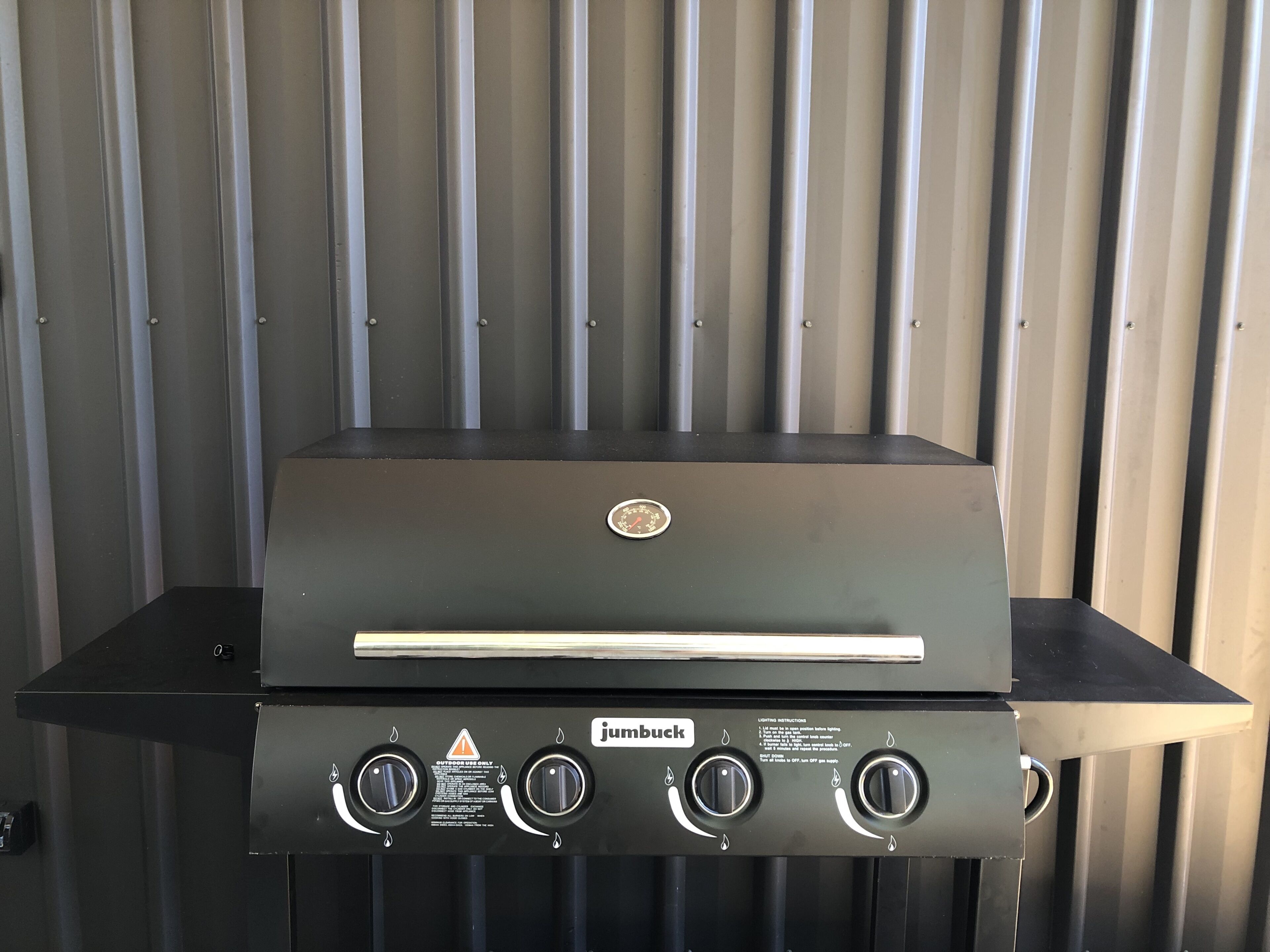 Een Jumbuck gasbarbecue voor buiten met vier branders en een bedieningspaneel, voor een golfplaten achtergrond.