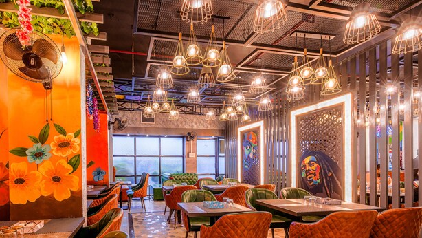 Un restaurant à l’ambiance chaleureuse, avec un intérieur décoré de couleurs vives, des chaises recouvertes d’un tissu vert et orange, un grand ventilateur et une fresque représentant des fleurs sur le mur.