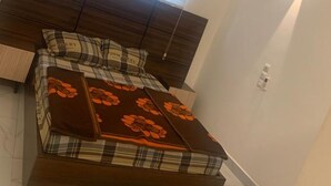 A bedroom with a dark wood headboard, a bed with a plaid bedspread and orange floral throw, a window with blinds, and a wall-mounted light fixture. - Logement Entier : Appartement 3 Pièce (Cotonou)