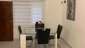 A dining area with a glass table and black chairs, a wooden door, a window with blinds, and a painting on the wall. - Logement Entier : Appartement 3 Pièce (Cotonou)