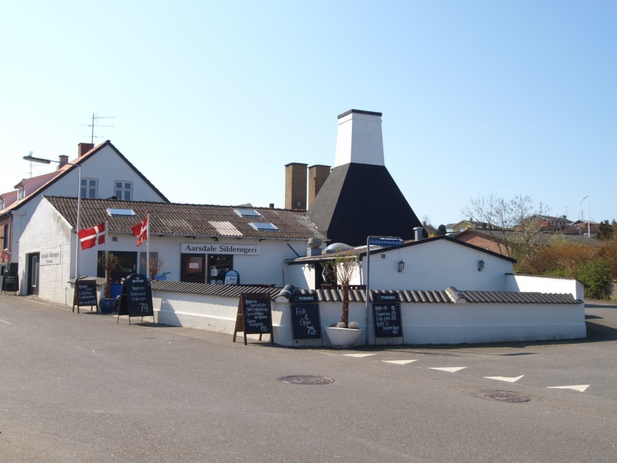 A building with a sign reading 'Aarsdale Silderogeri' and flags, a chimney, and outdoor seating area with chalkboard menus.