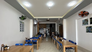Comedor de hotel con varias mesas y sillas azules, una escalera que conduce al piso superior y un cartel que indica las tarifas de las habitaciones.