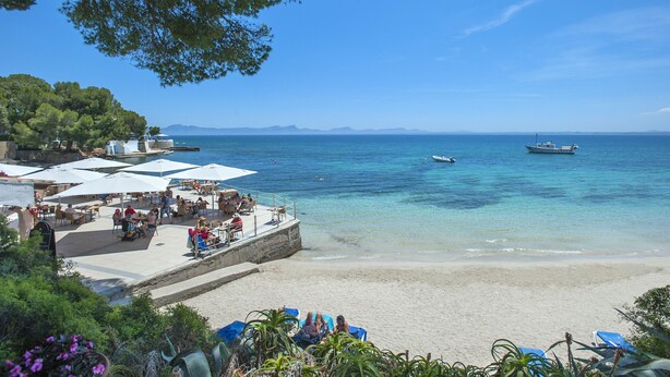 A beachside café with outdoor seating, a sandy beach, and a view of the sea and distant mountains. - VILLA GABRIEL - RUSTIC STAY (Alcúdia)