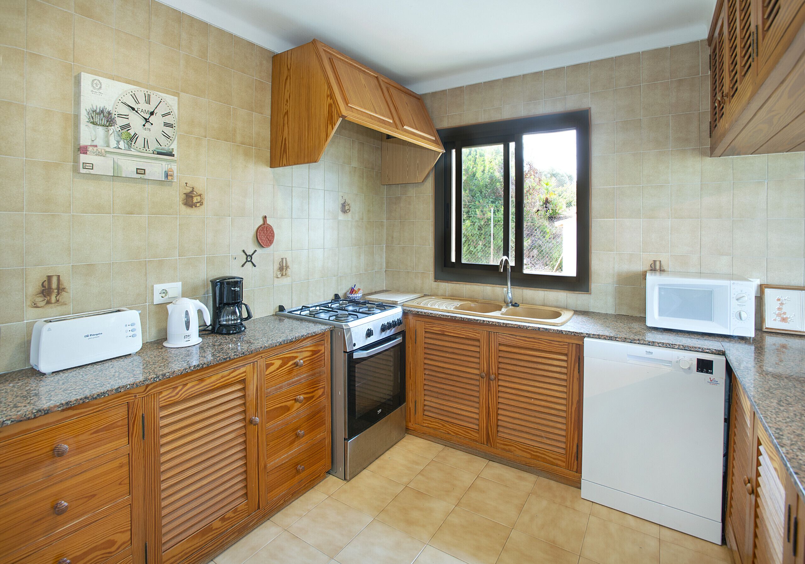 Cucina con pensili e basi in legno, fornello, lavastoviglie, microonde e una finestra.