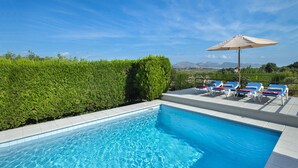 A swimming pool with clear blue water, surrounded by a low hedge, and a row of sun loungers with an umbrella. - VILLA GABRIEL - RUSTIC STAY (Alcúdia)