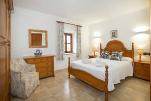 Un dormitorio con cama de madera, una mesita de noche de madera, un aparador de madera, una silla, una ventana con cortinas y un espejo instalado en la pared.