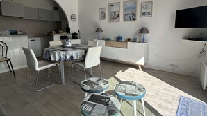Una cocina compacta con gabinetes blancos, un comedor con mesa y sillas, un televisor montado en la pared y una pequeña zona de estar con taburetes.