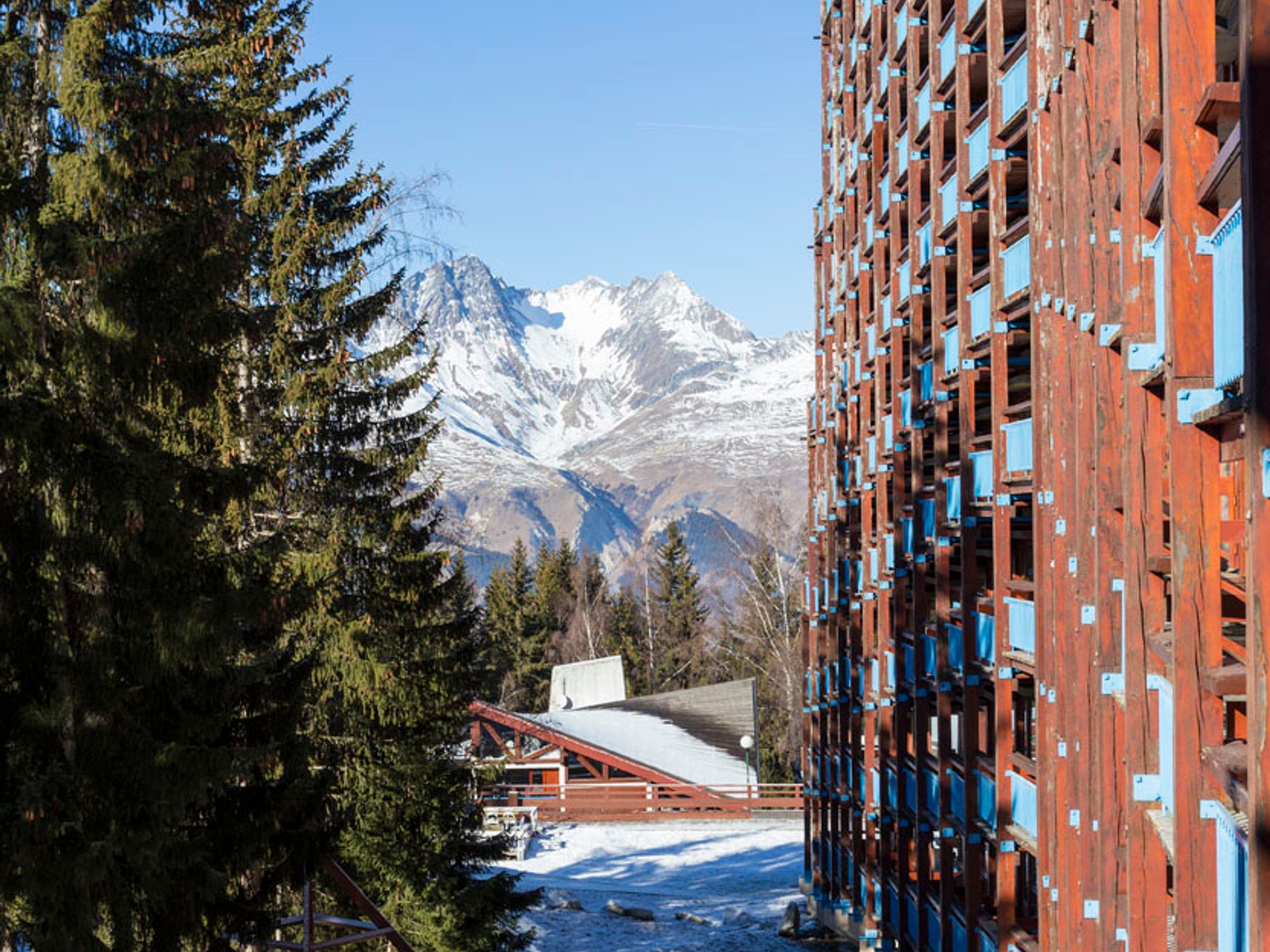 A building with a red facade and blue accents, a snow-covered mountain range in the background, and a forested area in the foreground.