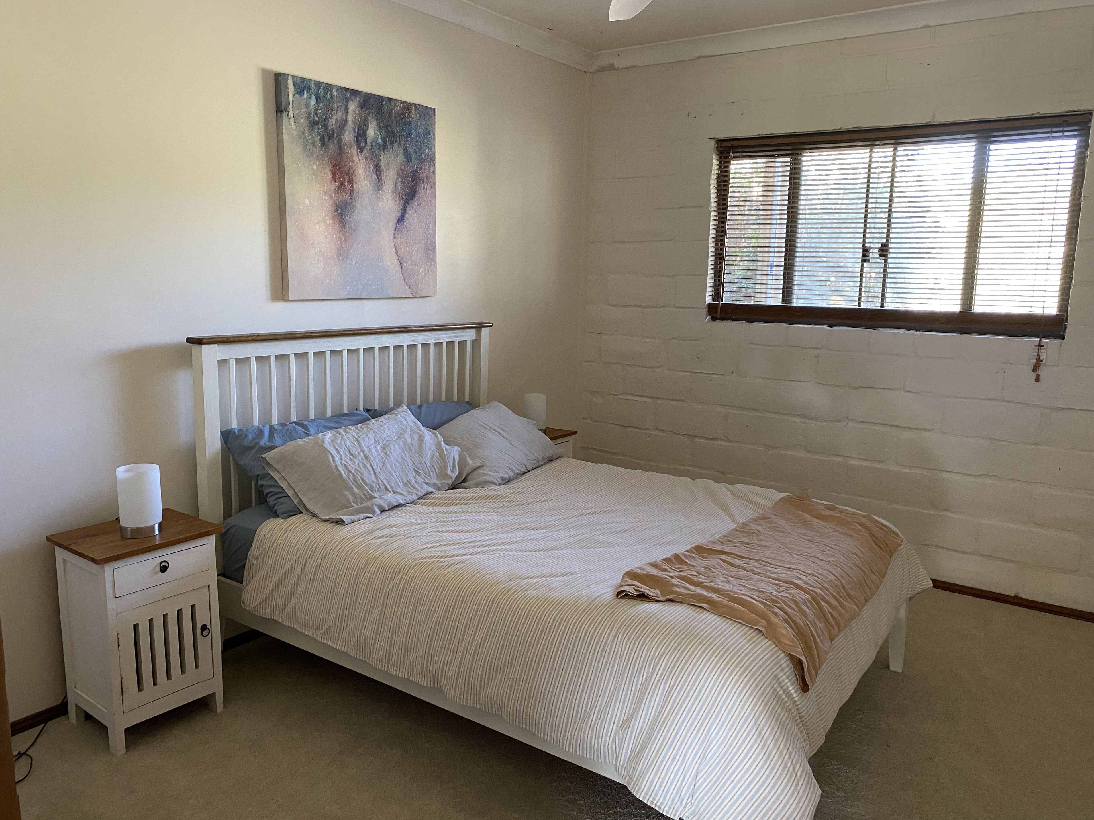 A bedroom with a bed, a nightstand, a bedside table, a window with blinds, and a wall painting.