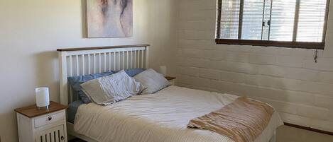 A bedroom with a bed, a nightstand, a bedside table, a window with blinds, and a wall painting.