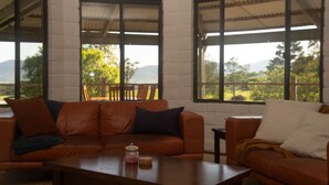 Una habitación con sofá de cuero, mesa de centro de madera y ventanas amplias con vistas a zonas verdes y montañas.