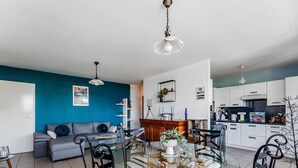 A modern living space with a blue accent wall, a glass dining table surrounded by striped chairs, a grey sofa, and a kitchen area with white cabinets and a blue countertop. - Biscarrosse beach, air-conditioned apartment with wifi, center, 4 pax (Biscarrosse Plage)