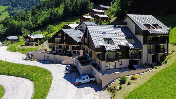 A modern multi-story building with a sloped roof, surrounded by lush greenery and a mountainous backdrop.