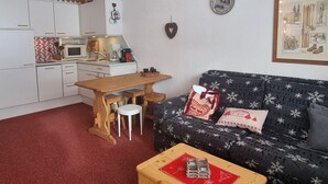 Ein kompakter Wohnraum mit Küchenzeile, einem Holzespeisetisch mit zwei Stühlen, einem schwarzen Sofa mit festlichen Kissen und einem roten Couchtisch.