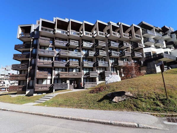 Un immeuble résidentiel à plusieurs étages, doté de balcons, d’un rez-de-chaussée en pierre et d’une pente herbeuse.