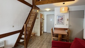 A wooden staircase leading to an upper level, a dining area with a table and chairs, and a red sofa in a room with wooden flooring and white walls.
