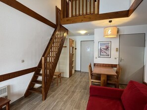 Uma escada de madeira leva a um andar superior, onde se encontra uma sala de jantar com mesa e cadeiras, e um sofá vermelho em um ambiente com piso de madeira e paredes brancas.
