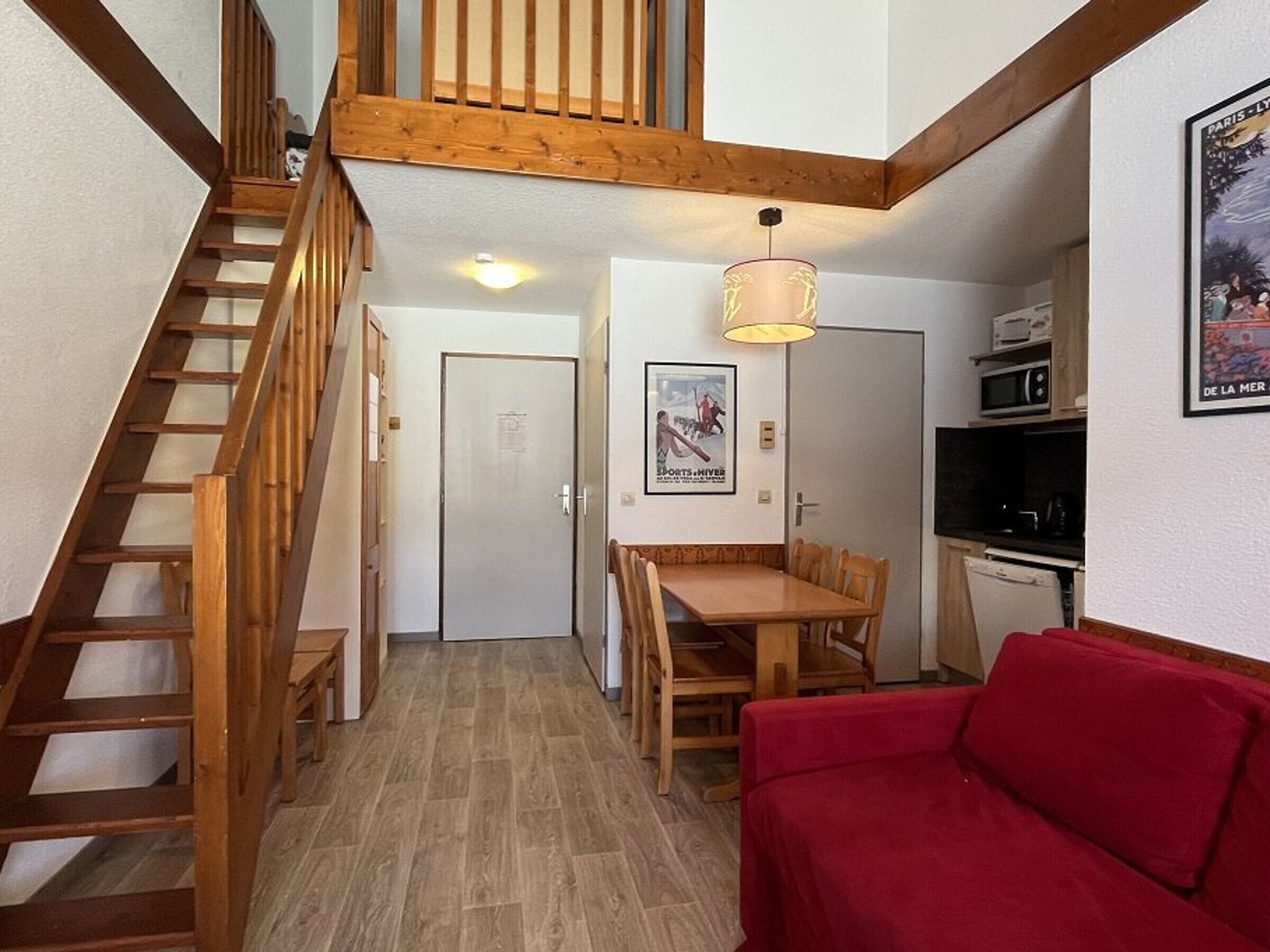 Un espacio habitable compacto con una escalera de madera, un área de comedor con mesa y sillas, un sofá rojo y una cocina con microondas.