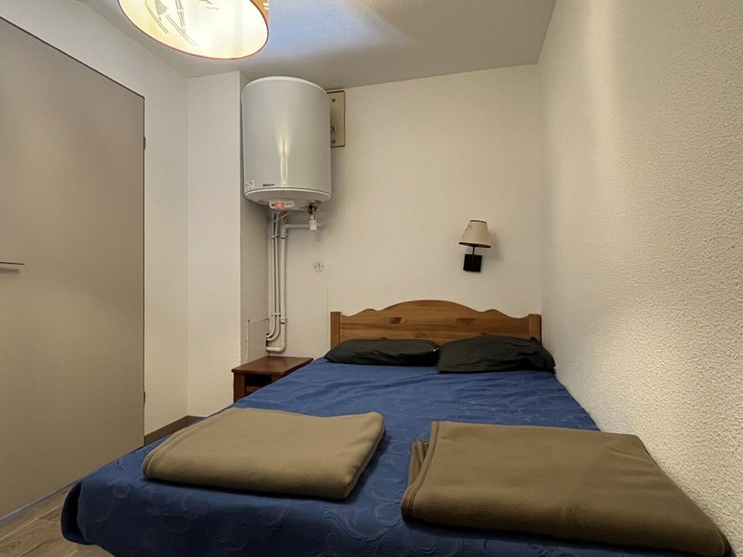 A bedroom with a bed, a nightstand, a wall-mounted heater, and a lamp.
