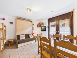 A room with a wooden bench, a small stool, a TV on the wall, and a wooden door with a window.