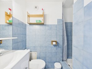A bathroom with blue tiles, a white toilet, a shower with a curtain, and wooden shelves with toiletries.
