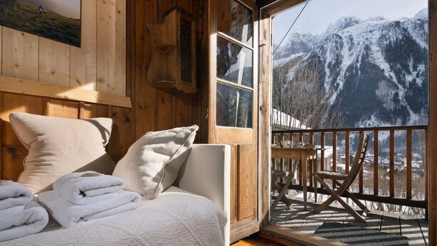 Une chambre rustique en bois, avec un lit, des oreillers et une vue sur des montagnes enneigées, visible à travers une porte vitrée.