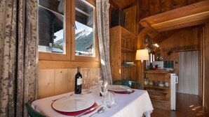Un intérieur en bois avec une table à manger pour deux personnes, une fenêtre donnant sur des montagnes enneigées et un coin cuisine équipé d’un réfrigérateur et d’étagères.