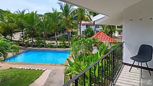 A balcony with a view of a swimming pool, surrounded by tropical plants and a red-roofed building. - Colorful Condo w/ Community Pool and Walking Distance from the Beach (Coco)