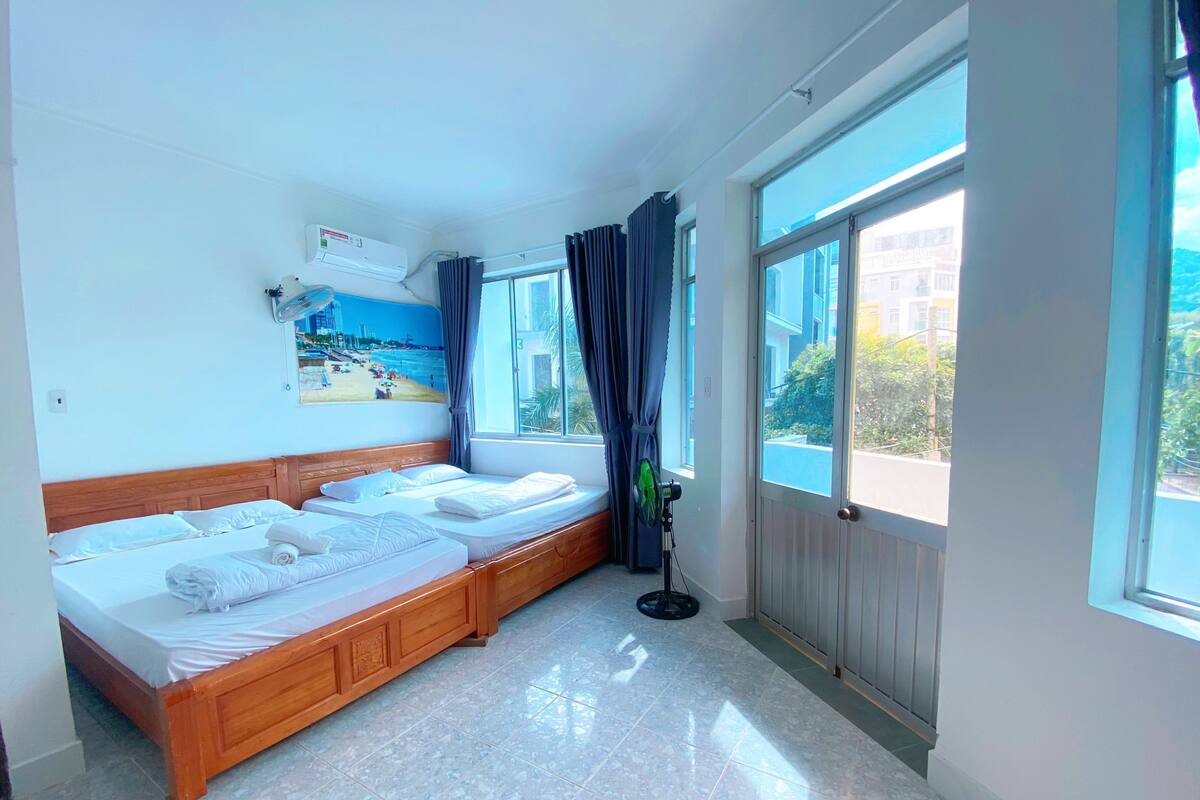 A hotel room with a wooden bed, a ceiling fan, a large window with curtains, and a view of the city.