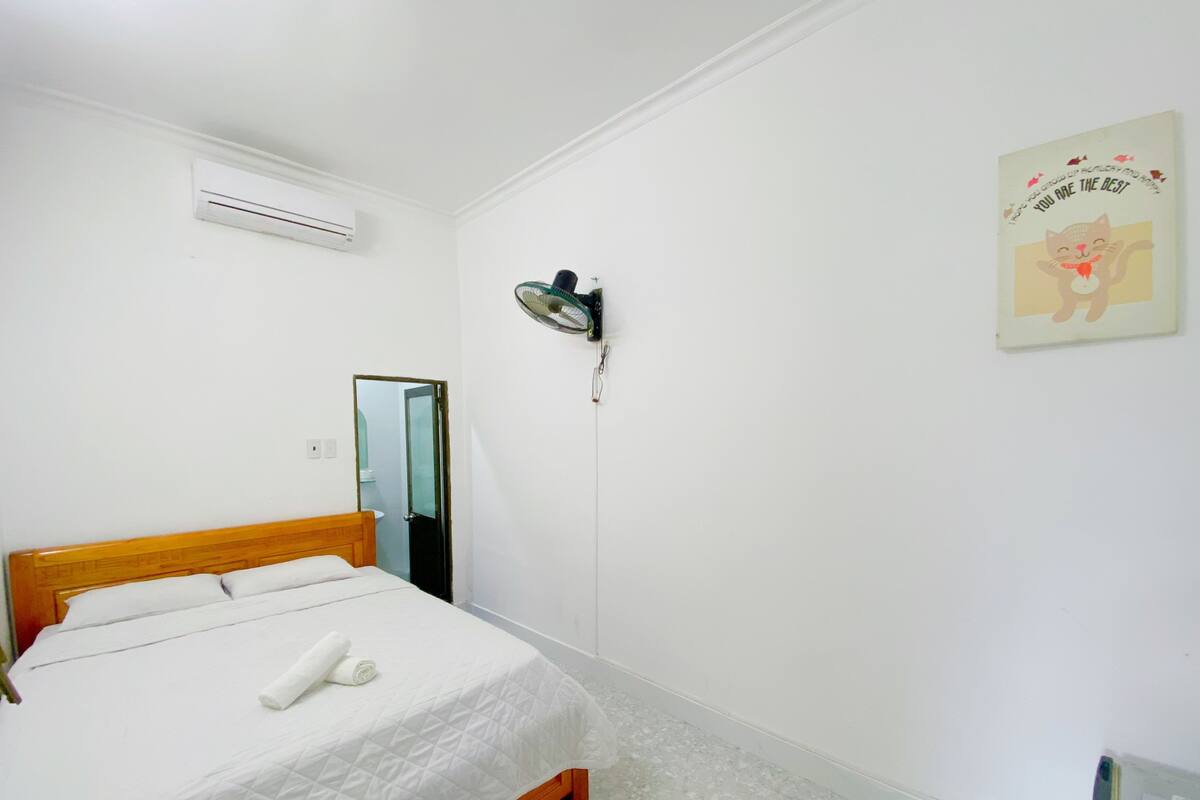 A neatly made bed with a wooden headboard, a wall-mounted air conditioner, a standing fan, a mirror, and a motivational poster on the wall.