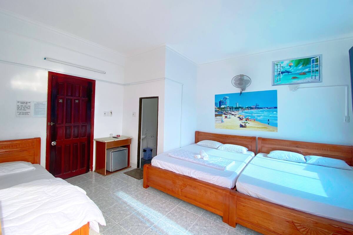 A hotel room with two single beds, a wooden headboard, a small desk with a chair, a mirror, a red door, and a beach-themed wall art.