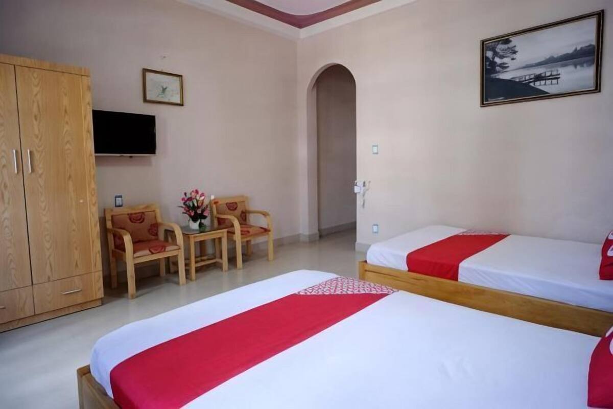 A hotel room with two beds, a TV, a wooden wardrobe, a desk with chairs, and a painting on the wall.