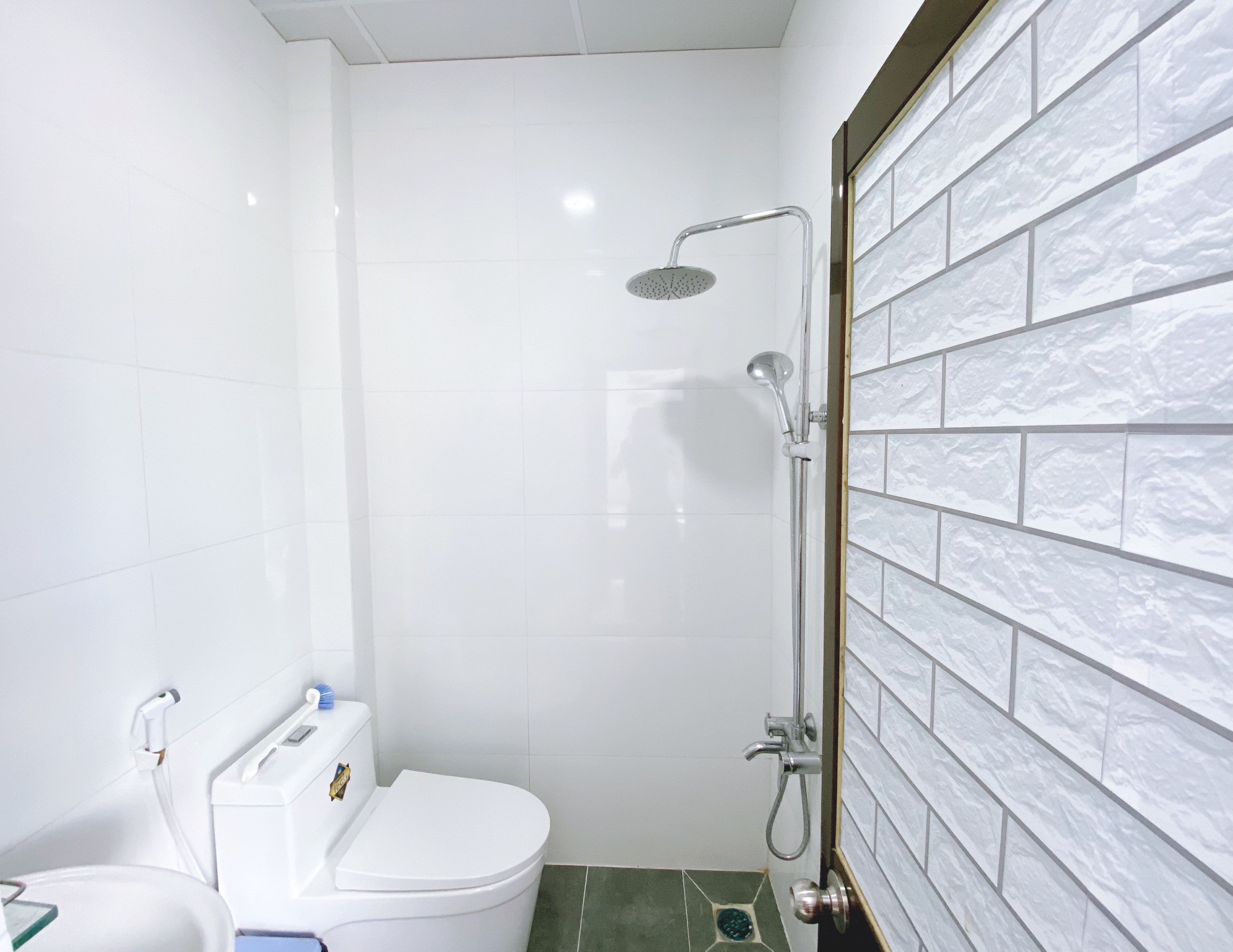 A small, modern bathroom with a white toilet, a shower with a handheld showerhead, and a tiled wall with a marble-like pattern.