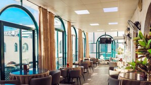 A restaurant with round tables and chairs, large windows with curtains, and a TV mounted on the wall. - AL YASMEEN HOTEL (Nablus)