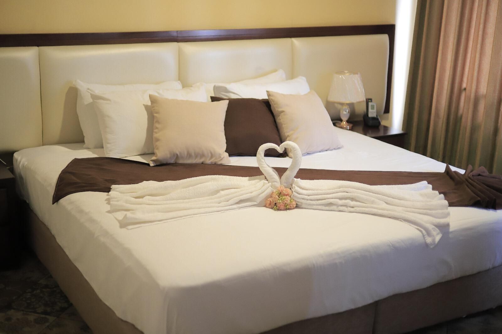 A hotel room with a neatly made bed, a heart-shaped towel arrangement, and a nightstand with a lamp.