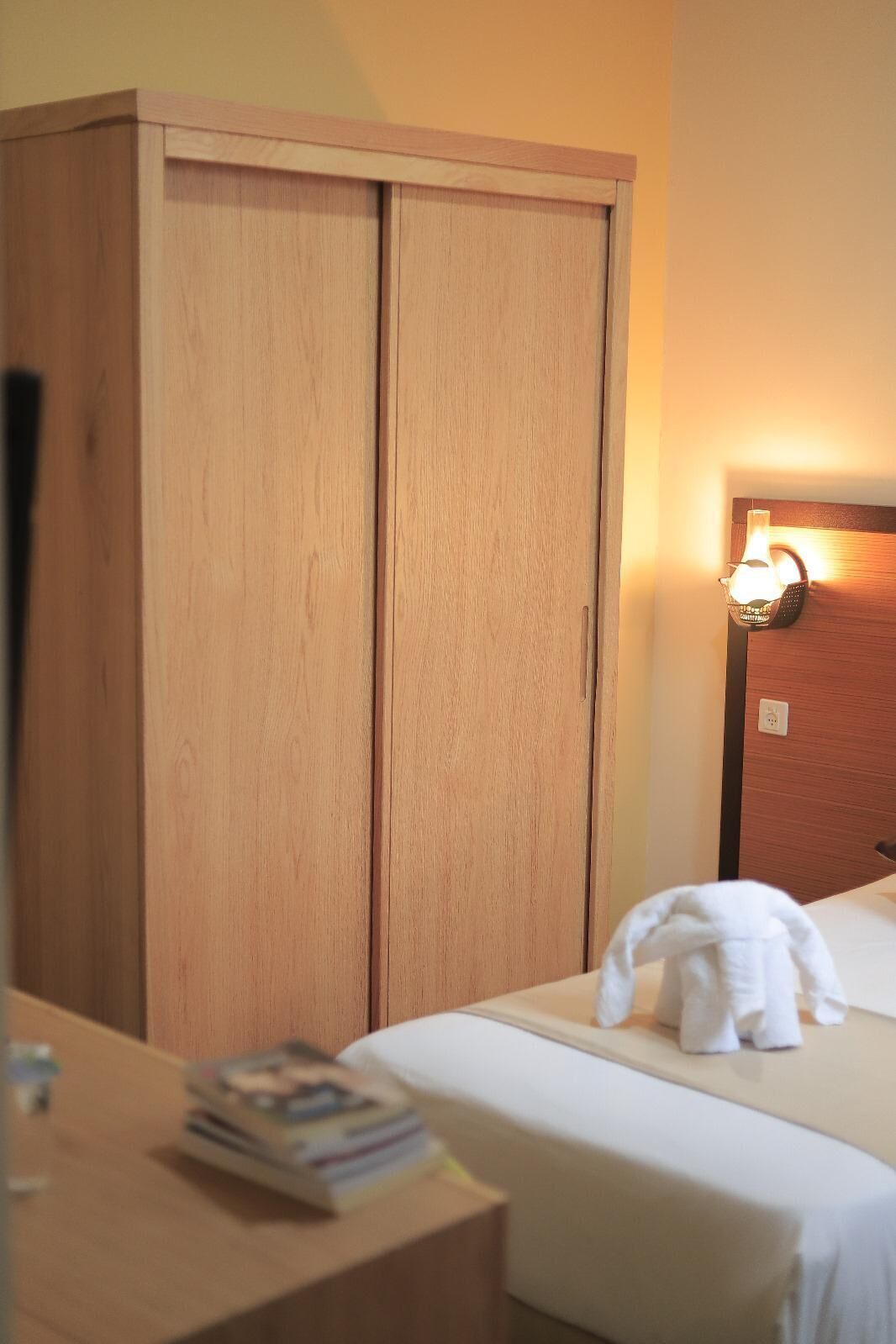 A hotel room with a wooden wardrobe, a bedside lamp, and a neatly made bed with a white towel.