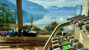 A view of a lake with mountains in the background, a window with exercise equipment, and a clear sky. - Ideal Lakeside Retreat: 2-bedroom Spa ap. With Lake View, Centre, Sleeps 1-4 (Montreux)