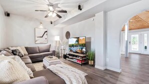 A modern living room with a sectional sofa, a flat-screen TV, a ceiling fan, and a large window.