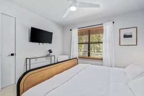 A bedroom with a bed, a TV mounted on the wall, a desk with a chair, and a window with curtains.